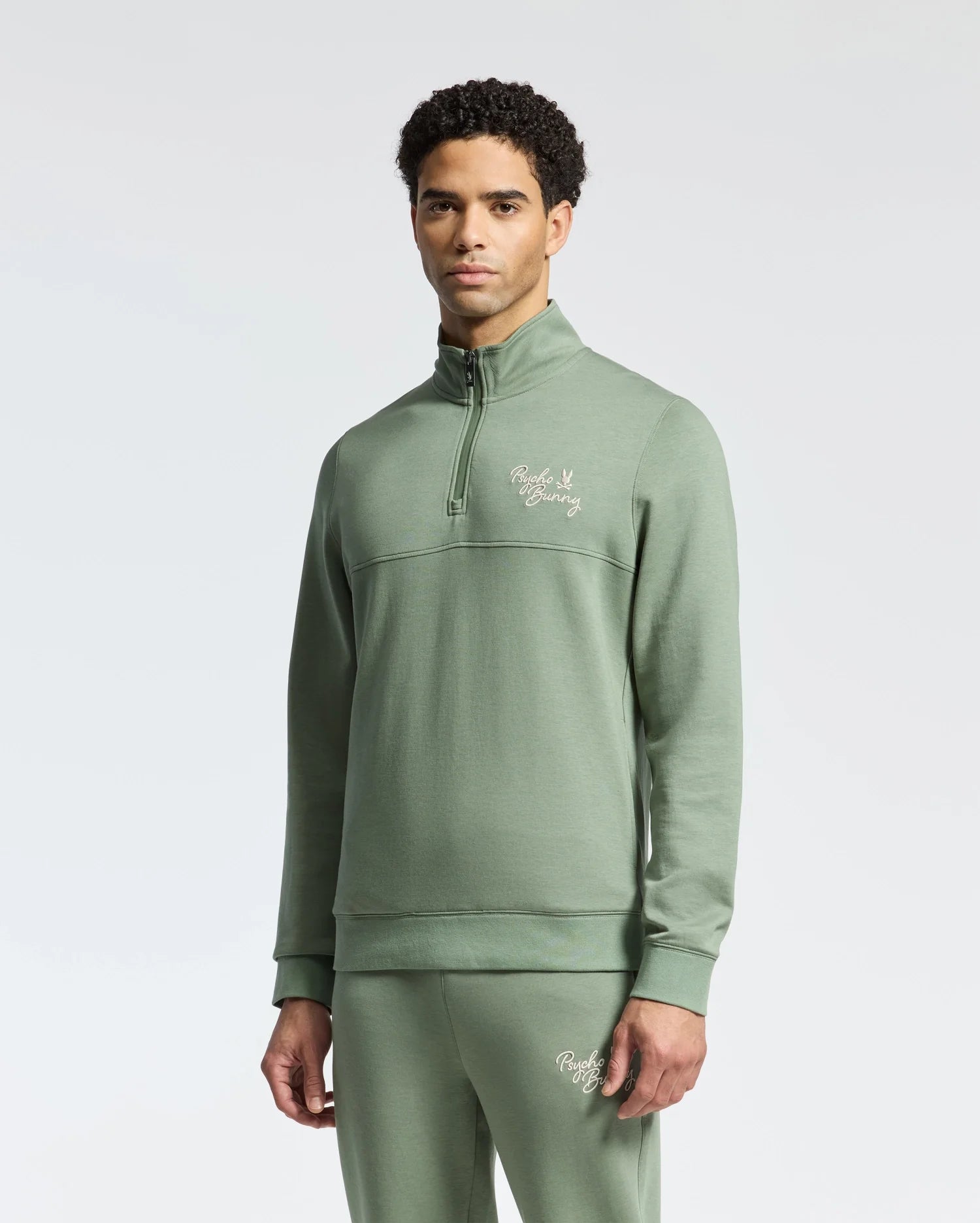 A man in the Psycho Bunny MENS DESERT 1/4 MOCK ZIP - B6S488F200, a light green 100% cotton quarter-zip pullover with matching pants, both with an embroidered wordmark logo on the chest and thigh, stands against a plain white background.