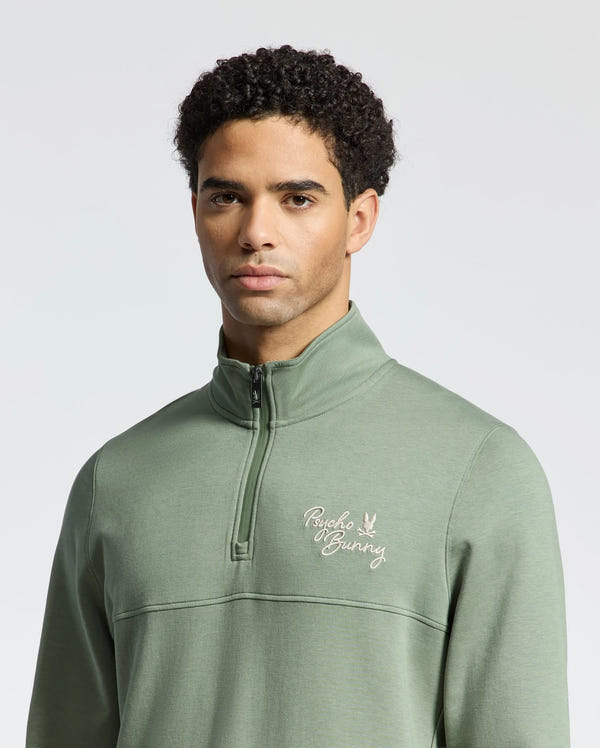 A man wears the "MENS DESERT 1/4 MOCK ZIP - B6S488F200" sweatshirt in sage green, made of 100% cotton, with a "Psycho Bunny" wordmark logo on the left chest. A light gray background complements its chic casual style.