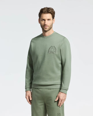 A man wears a Psycho Bunny Melbourne Crewneck in sage green, made from breathable Modal blend, paired with matching pants. The soft stretch jersey sweatshirt features a subtle chest logo, standing out against the plain white background.