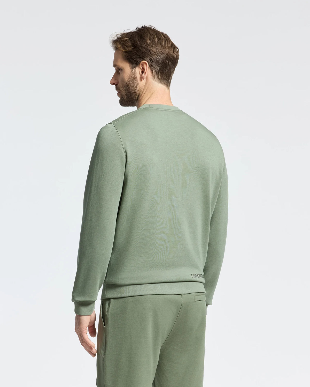 A man stands against a plain backdrop in a sage green Psycho Bunny Melbourne Embroidered Classic Modal Crewneck Sweatshirt, paired with matching pants. Facing sideways, he looks left with short hair and a beard, embodying relaxed elegance.