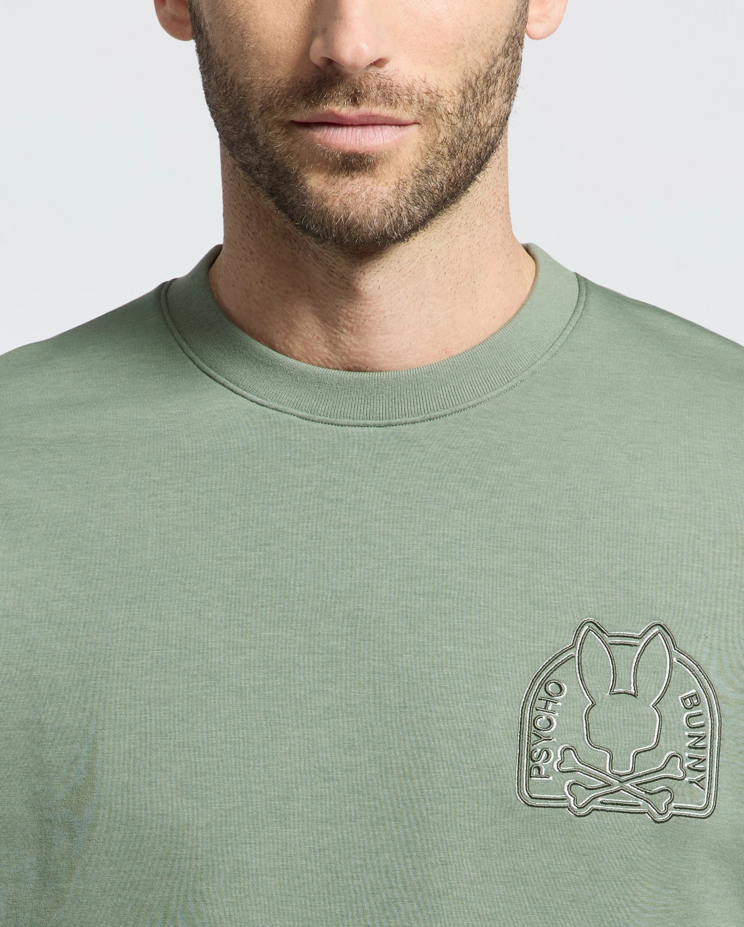A person wearing the Psycho Bunny Men's Melbourne Embroidered Classic Modal Crewneck Sweatshirt, featuring a stylized bunny above crossed bones with "Psycho Bunny" text in a circular logo on the chest, all embossed in a slightly lighter green on soft stretch jersey fabric.