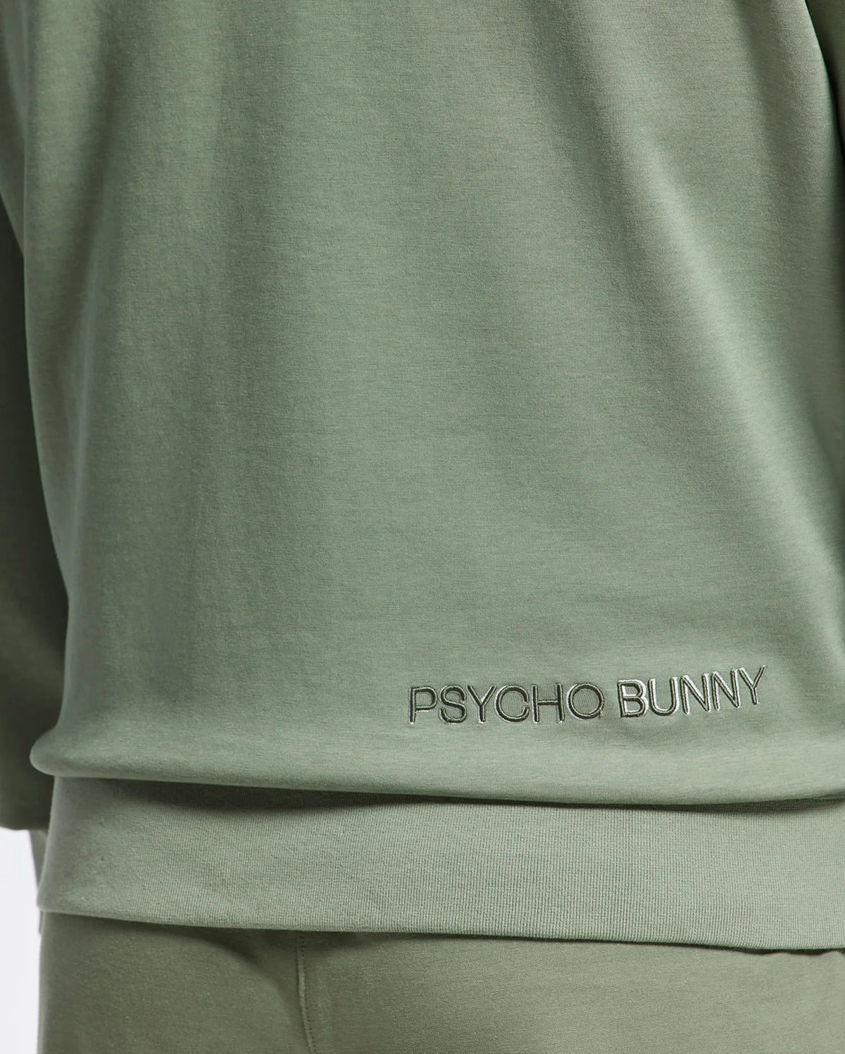 Close-up of a person wearing a Psycho Bunny MELBOURNE Crewneck in light green with lower back embroidery, crafted from a smooth Modal blend. Matching green pants are partially visible below.