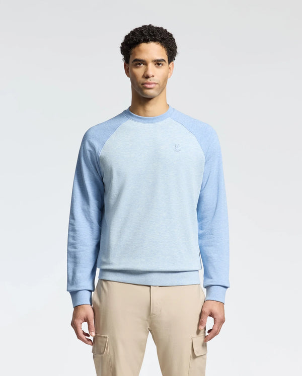 A man stands against a neutral background wearing a Psycho Bunny MENS ARCHIE MELANGE COLOR BLOCK SWEATSHIRT in light blue with darker blue sleeves and an embroidered Bunny, paired with beige pants. His hands are by his sides.