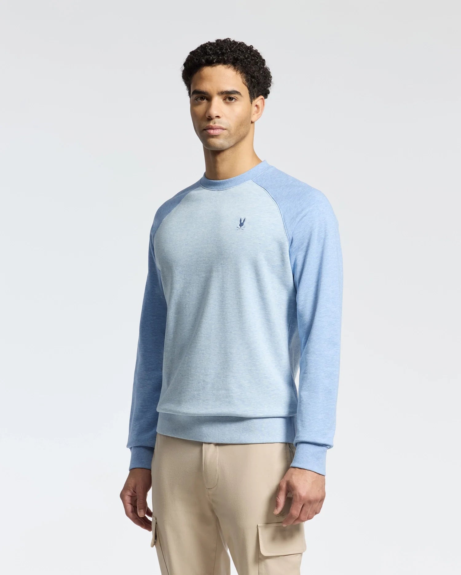 A man in a Psycho Bunny MENS ARCHIE MELANGE COLOR BLOCK SWEATSHIRT (B6S630F200) with light blue and darker blue raglan sleeves, plus beige cargo pants, stands against a white background. The sweater has an embroidered Bunny logo on the chest, adding a playful touch to his outfit.