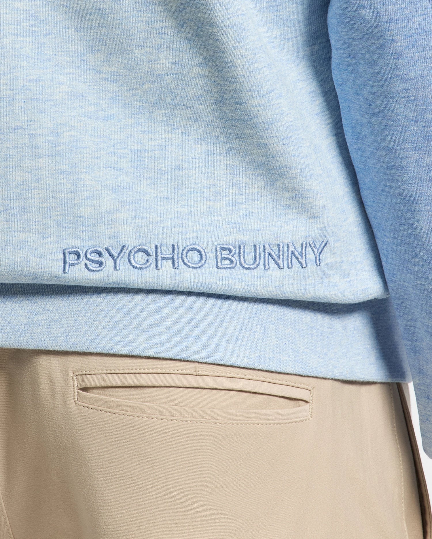 Close-up of a person wearing the Psycho Bunny MENS ARCHIE MELANGE COLOR BLOCK SWEATSHIRT, B6S630F200, in light blue with an embroidered Bunny logo at the hem, paired seamlessly with beige pants for a subtle monochrome styling.