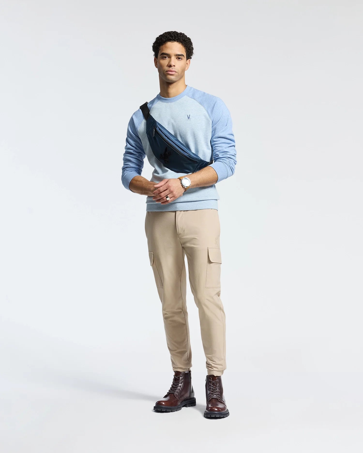 A man poses against a plain backdrop in monochrome style, wearing a light blue Psycho Bunny ARCHIE melange color block sweatshirt, beige cargo pants, and brown boots. His black crossbody bag features an embroidered Bunny, and he wears a watch on his left wrist. He gazes ahead with ease.
