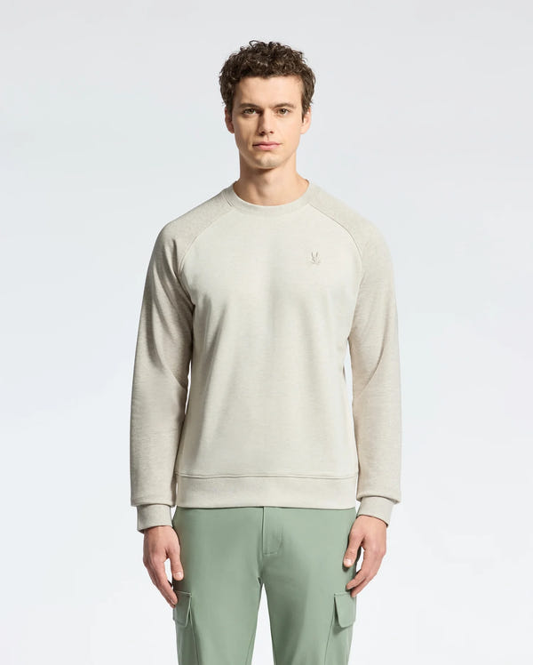 A person stands against a white background wearing a Psycho Bunny MENS ARCHIE MELANGE COLOR BLOCK SWEATSHIRT, featuring an embroidered bunny emblem. Paired with green cargo pants, the outfit complements their short curly hair in a monochrome style.