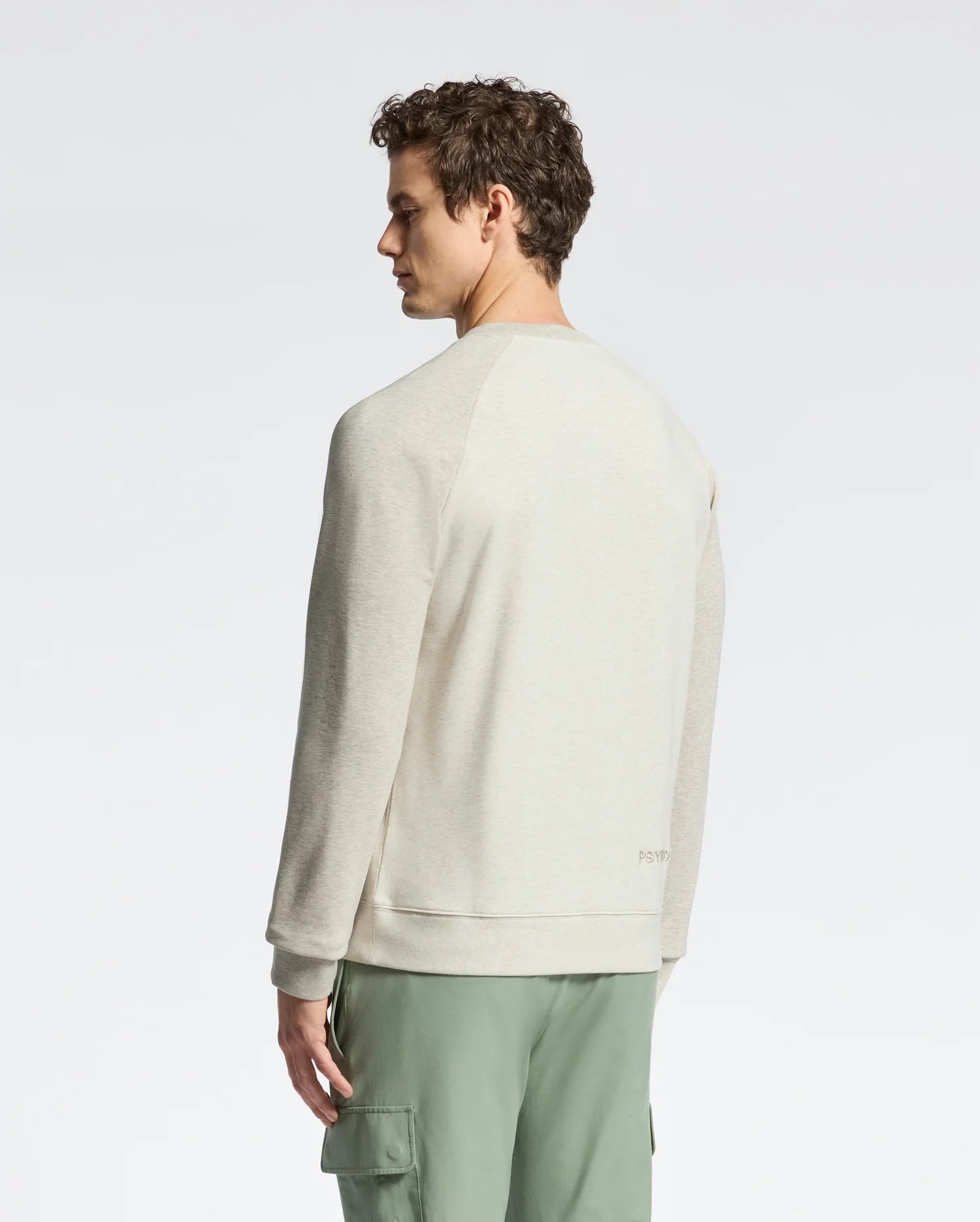A person in a Psycho Bunny MENS ARCHIE MELANGE COLOR BLOCK SWEATSHIRT - B6S630F200 stands sideways, wearing green cargo pants and gazing down. The simple studio backdrop complements the outfit's understated elegance.