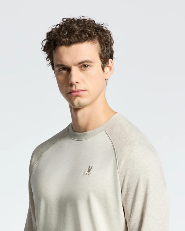 A person with curly hair, wearing Psycho Bunny's light gray Archie Melange Color Block Sweatshirt, made of soft cotton jersey and featuring a small embroidered bunny logo on the chest, stands against a plain, light background.