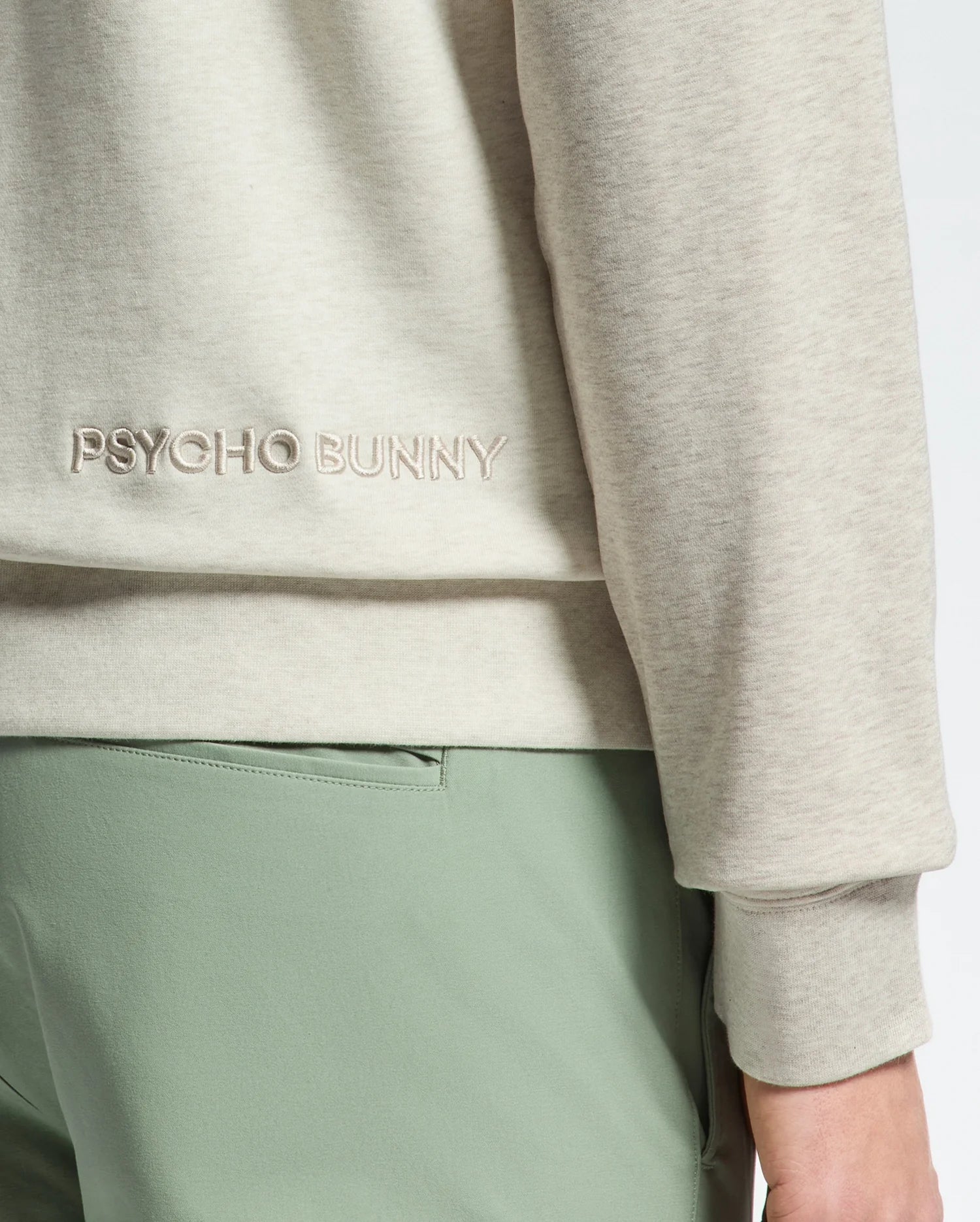 A person wears the ARCHIE MELANGE COLOR BLOCK SWEATSHIRT by Psycho Bunny, featuring a beige cotton design with "PSYCHO BUNNY" and an embroidered bunny. The sleeve complements their light green pants against a plain backdrop.