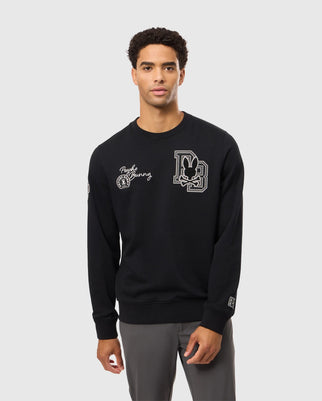 A man is showcasing the Psycho Bunny MENS ZACK VARSITY SWEATSHIRT - B6S880D200, embellished with chenille embroidery patches featuring a vintage-inspired logo. He poses against a plain gray background, pairing it seamlessly with gray pants.