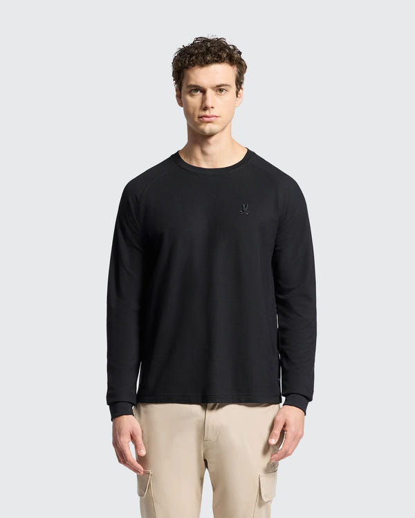 Against a light gray background, a person with curly hair wears beige cargo pants and the Psycho Bunny MENS TAYLOR PIQUE LONG SLEEVE TEE in black, featuring a small, subtle logo on the chest.