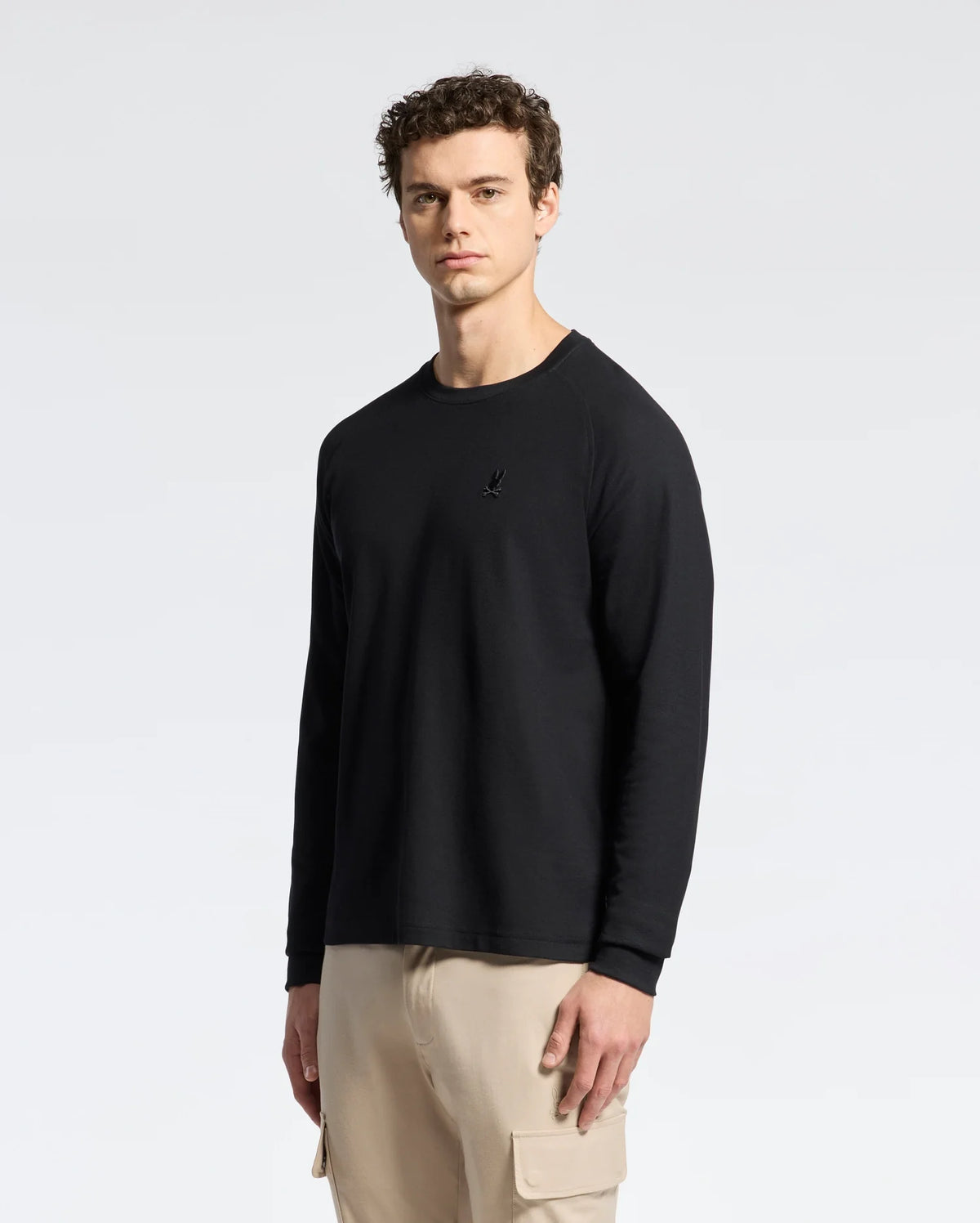 A person with curly hair stands against a plain white background, wearing a Psycho Bunny MENS TAYLOR PIQUE LONG SLEEVE TEE and beige cargo pants. Their neutral expression seamlessly combines casual comfort with understated style.