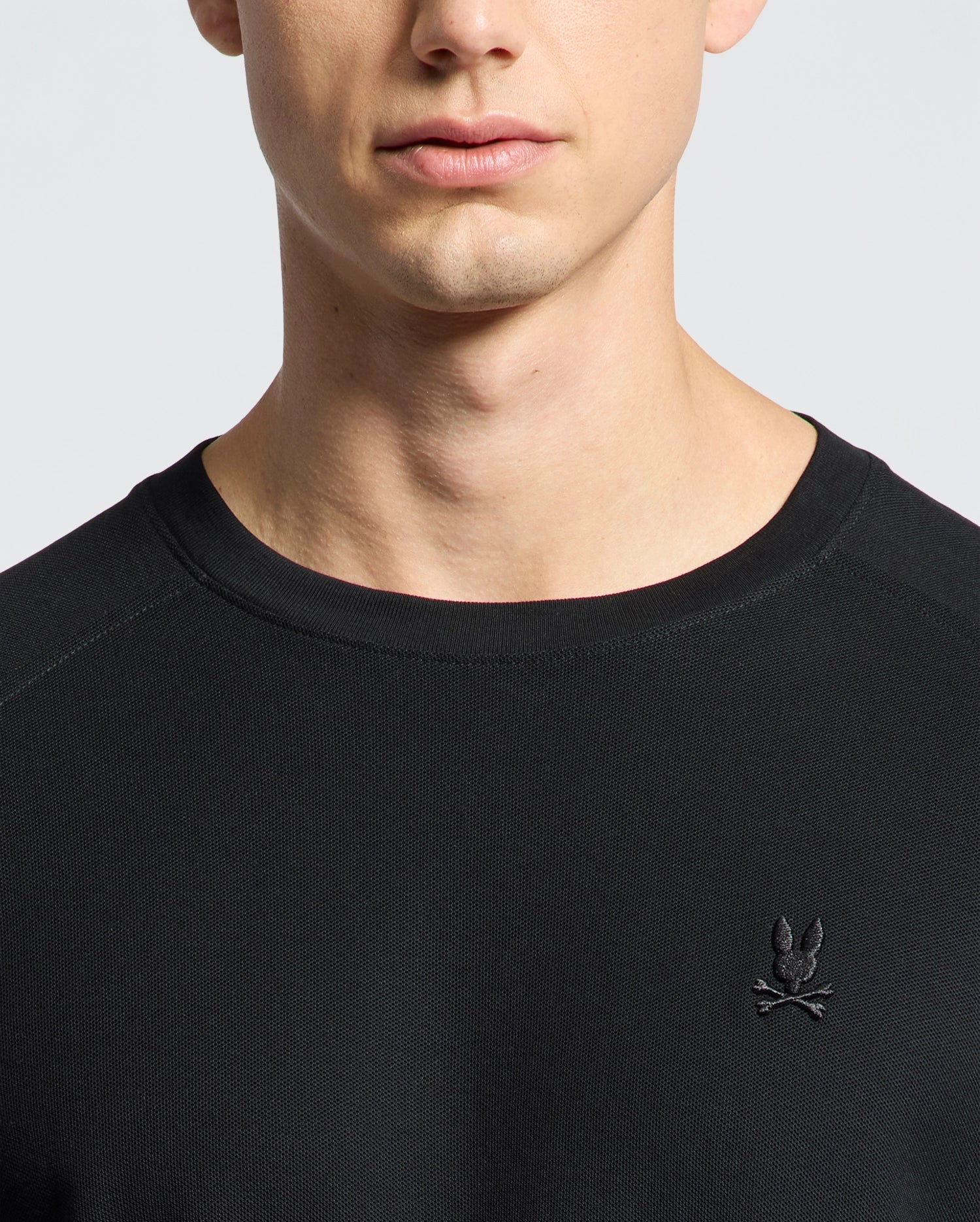 The MENS TAYLOR PIQUE LONG SLEEVE TEE by Psycho Bunny is a sleek black sweater crafted from soft Pima cotton. It features an embroidered rabbit and crossbones logo on the chest, set against a plain white background to highlight its luxurious texture.