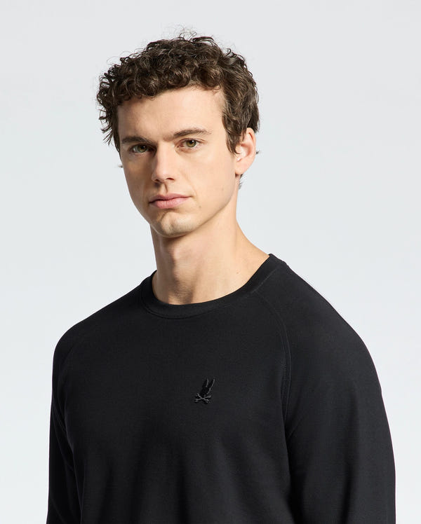 A person with curly brown hair is wearing the Psycho Bunny MENS TAYLOR PIQUE LONG SLEEVE TEE - B6T580F200, featuring a small embroidered emblem on the chest. The background is plain and light-colored.