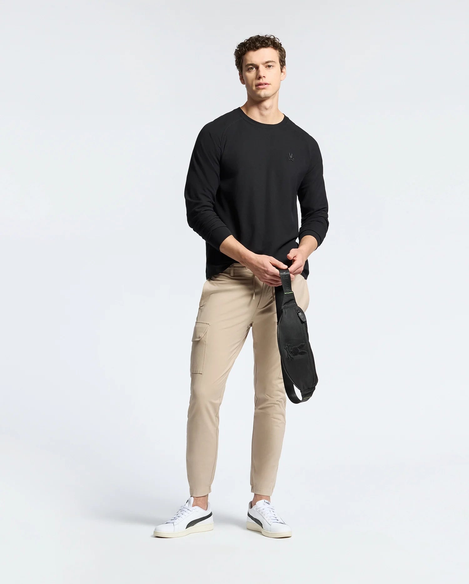 A man stands against a plain background wearing a Psycho Bunny MENS TAYLOR PIQUE LONG SLEEVE TEE in black, beige cargo pants, and white sneakers. He holds a black crossbody bag in front of him.