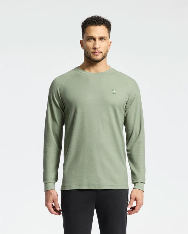 A man stands facing forward in a Psycho Bunny olive MENS TAYLOR PIQUE LONG SLEEVE TEE - B6T580F200 with an embroidered Bunny logo on the left chest, paired with black pants, set against a plain white background.