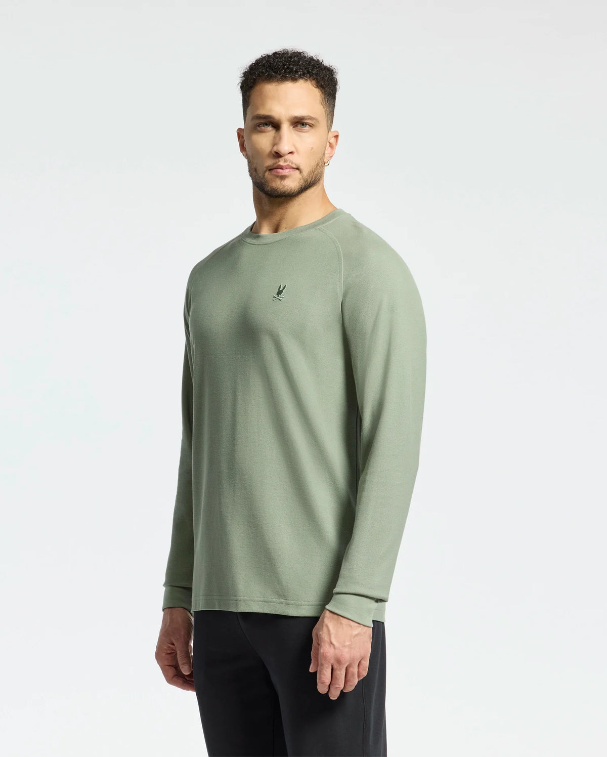 A man with short curly hair, dressed in a sage green Psycho Bunny MENS TAYLOR PIQUE LONG SLEEVE TEE and black pants, stands against a light gray background, looking directly forward.