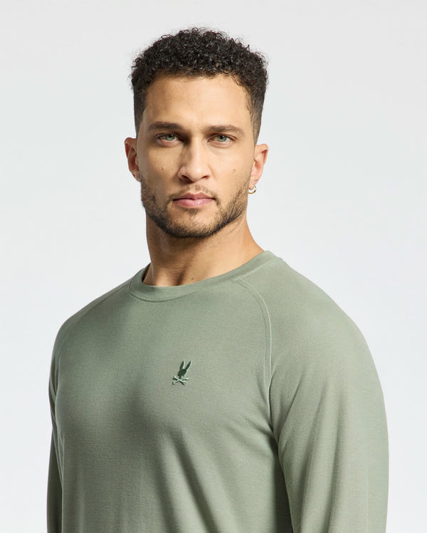 A person with short curly hair, a beard, and an earring wears a sage green Psycho Bunny MENS TAYLOR PIQUE LONG SLEEVE TEE featuring an embroidered bunny logo. They are looking directly at the camera against a plain, light-colored background.
