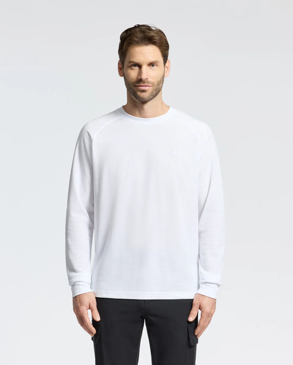 A person with short brown hair and a beard stands against a plain background, wearing the Psycho Bunny Men's Taylor Pique Long Sleeve Tee in white, paired with black pants, and looks directly at the camera with a neutral expression.