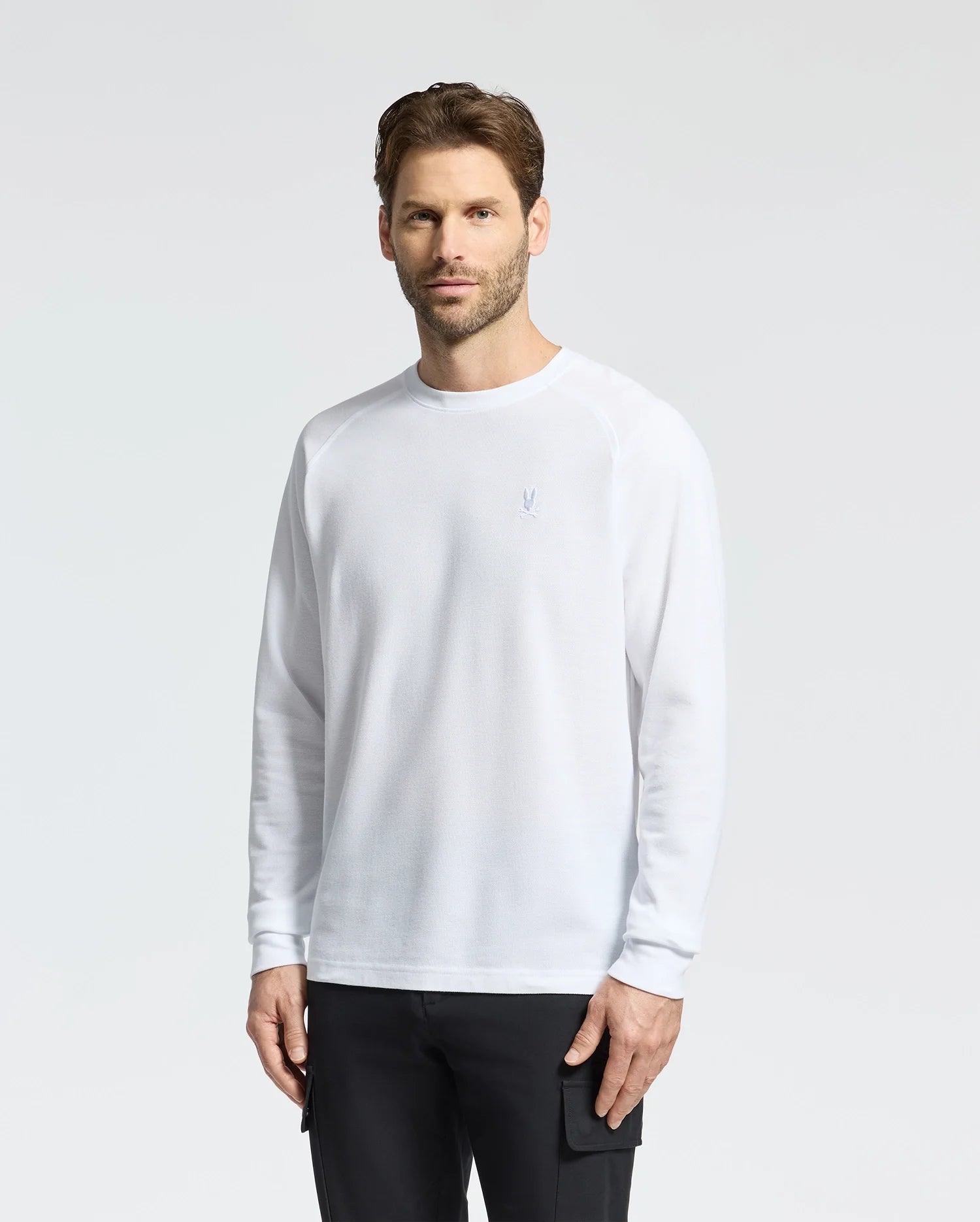 A man with short brown hair and a beard stands against a plain backdrop wearing black pants and the Psycho Bunny "Mens Taylor Pique Long Sleeve Tee" (B6T580F200), crafted from white piqué cotton with an embroidered bunny logo on the left chest.
