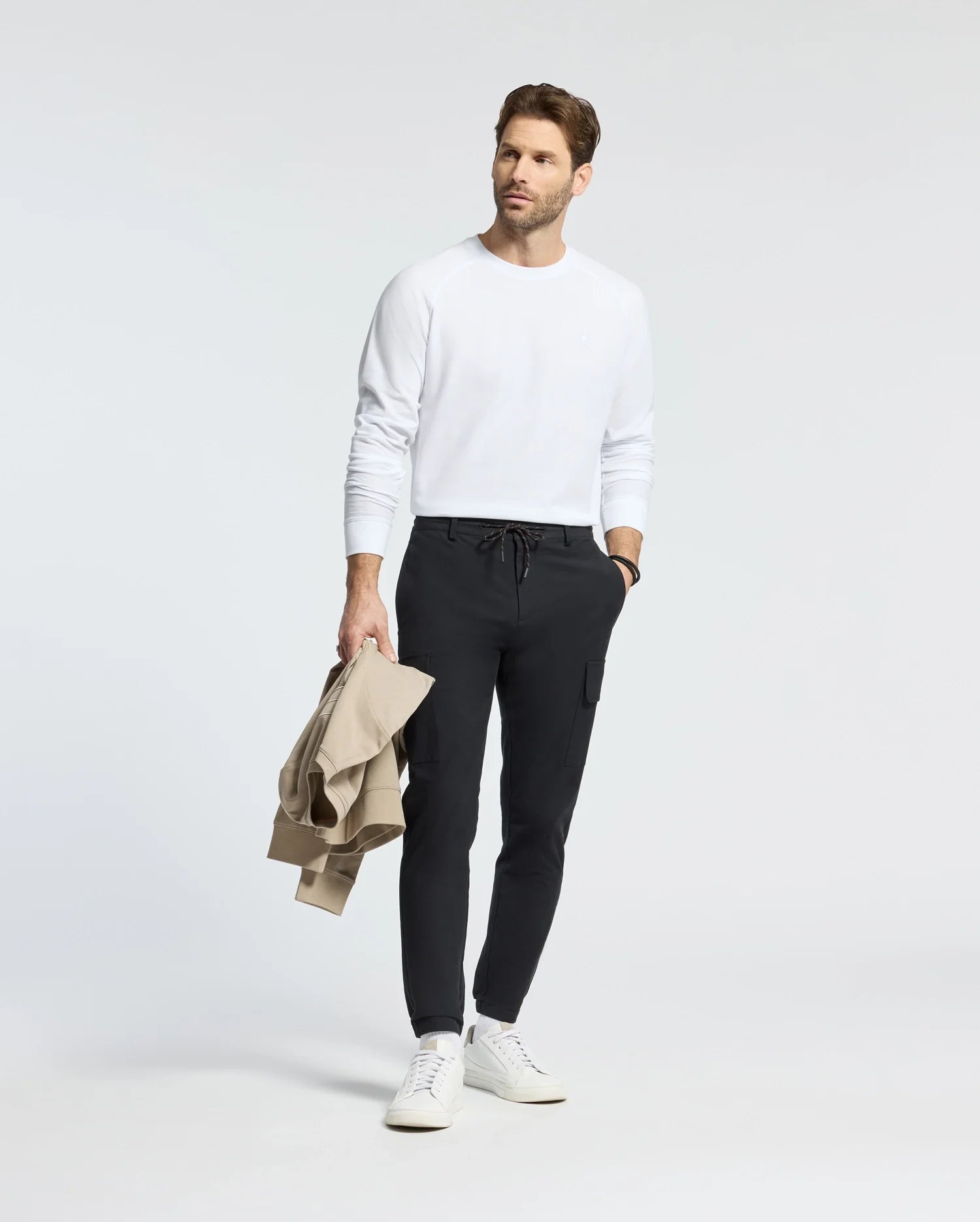 A man in a Psycho Bunny white Taylor Pique long sleeve tee and black jogger pants holds a beige jacket. He wears white sneakers, standing stylishly against a plain background with understated charm.