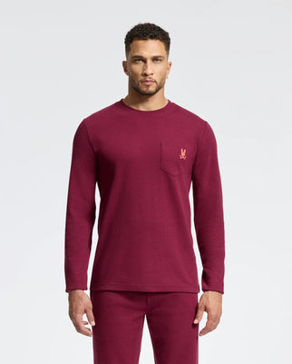 A person stands forward wearing a Psycho Bunny men's Val waffle lounge long sleeve tee and matching burgundy pants. The top features a small embroidered logo on the chest pocket, combining comfort with style against a plain white background.
