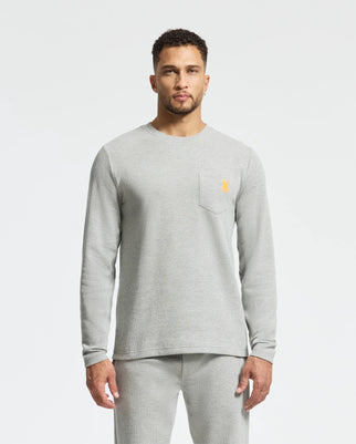 A man in a Psycho Bunny light gray long-sleeve tee, crafted from Pima cotton blend, stands against a plain white background. The MENS VAL WAFFLE LOUNGE LONG SLEEVE TEE features a small yellow logo on the chest pocket and pairs effortlessly with matching pants for a stylish look.