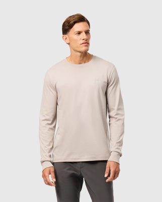 A person wearing a light grey Psycho Bunny MENS ALBERTON LONG SLEEVE TEE - B6T867D200 made from luxurious Pima cotton and dark grey pants stands against a plain background. The shirt, a fashion masterpiece, boasts a small logo on the left chest area. The person has short brown hair and is looking slightly to the left.