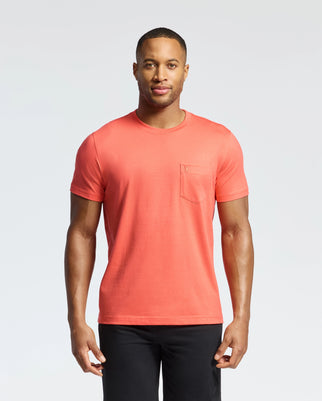 A man stands facing forward in a coral MENS BONNIE REGULAR FIT POCKET TEE by Psycho Bunny, made of soft Pima cotton featuring a chest pocket, paired with black pants against a plain white background.