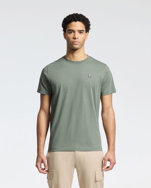 A man stands against a plain backdrop wearing Psycho Bunny's MENS CLASSIC CREW NECK TEE - B6U014B200 in green, crafted from soft Pima cotton and featuring a small left-chest logo. He pairs it with beige cargo pants, while his short curly hair and neutral expression complete the look.