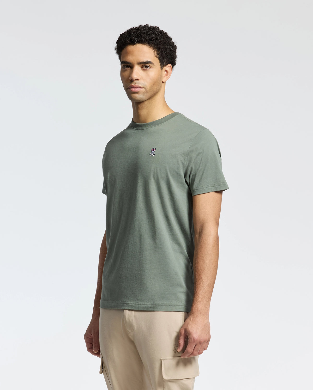 A man showcases the Psycho Bunny MENS CLASSIC CREW NECK TEE, made from Pima cotton in green with an embroidered logo, paired with beige cargo pants on a white background, delivering a relaxed regular fit.