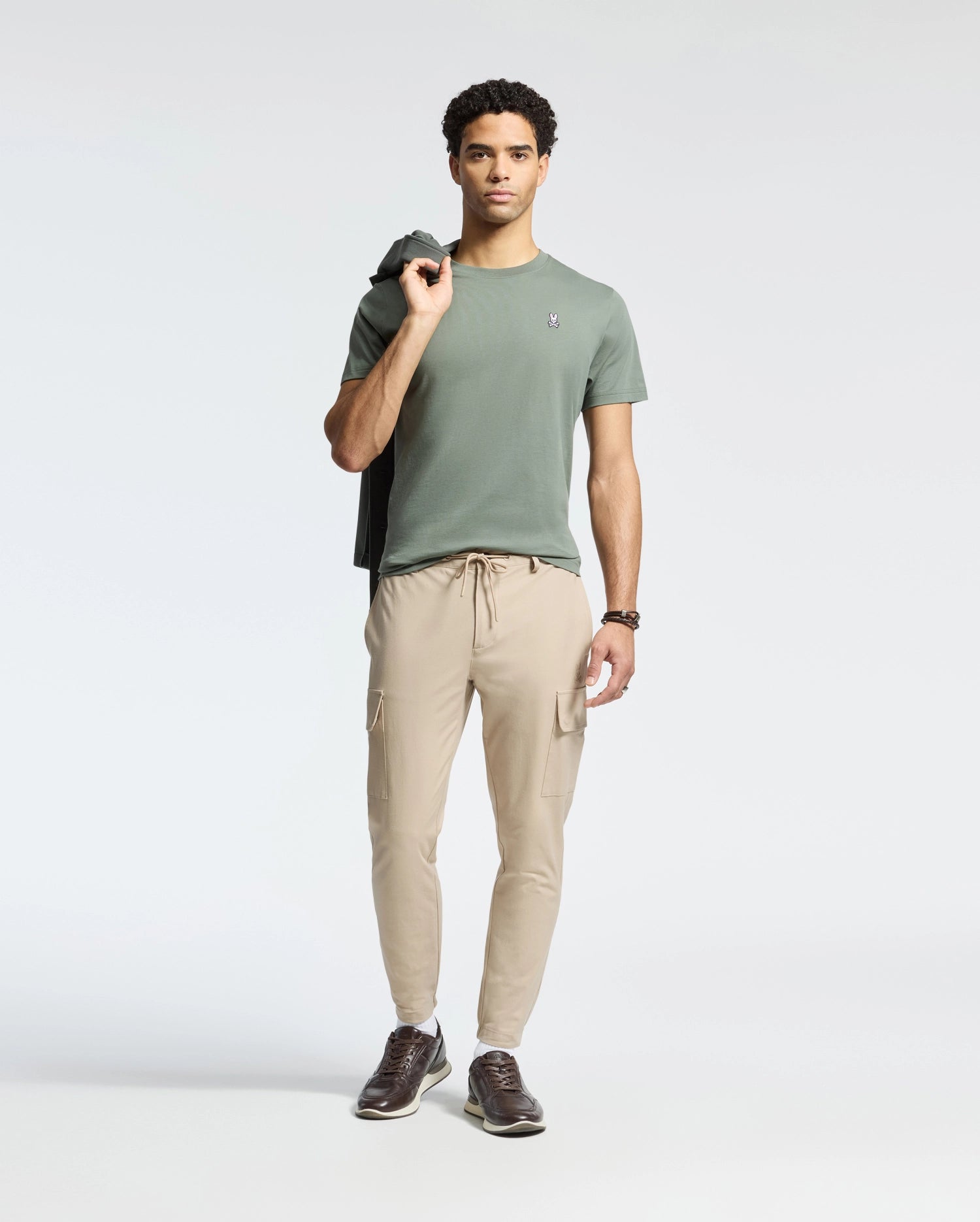 A person poses with a jacket over one shoulder, wearing the Psycho Bunny MENS CLASSIC CREW NECK TEE in green Pima cotton, paired with beige cargo pants and brown shoes. The plain white background highlights their relaxed stance and regular fit style.