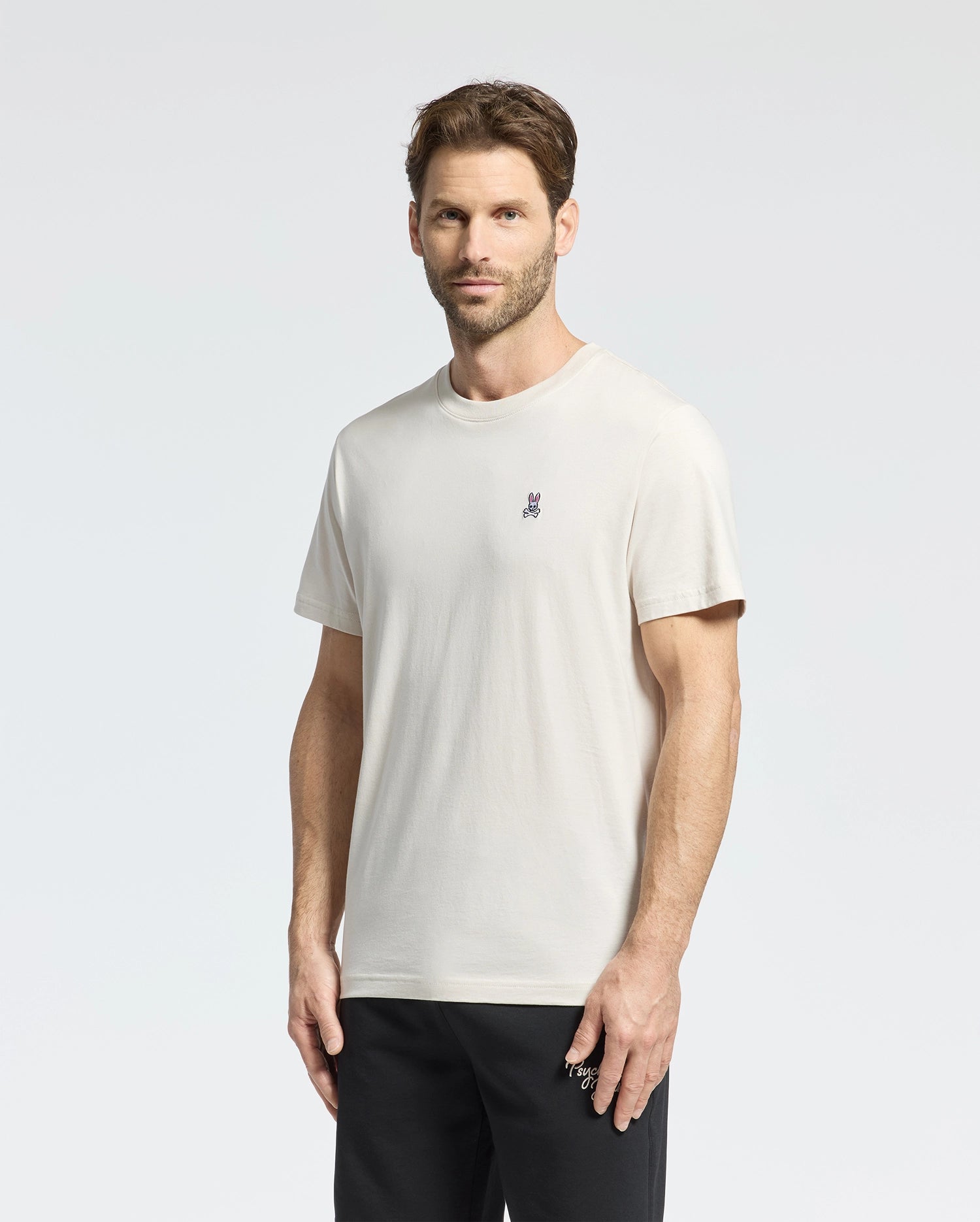 A man with short brown hair wears a Psycho Bunny Men's Classic Crew Neck Tee in premium jersey with a small embroidered chest logo, paired with black pants. He stands against a plain white background, looking at the camera neutrally.