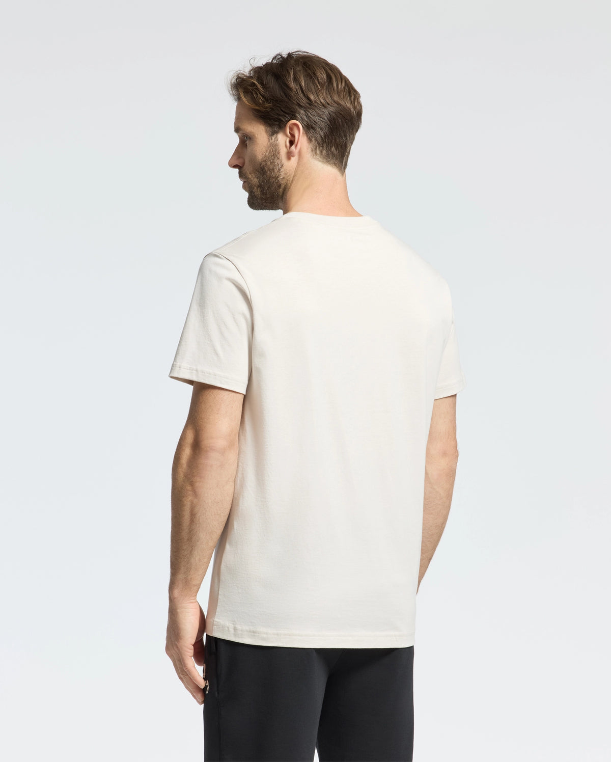 A man with short brown hair is seen from behind, wearing a Psycho Bunny MENS CLASSIC CREW NECK TEE - B6U014B200 made of premium jersey. His plain white T-shirt contrasts subtly against the light-colored background alongside his black pants.