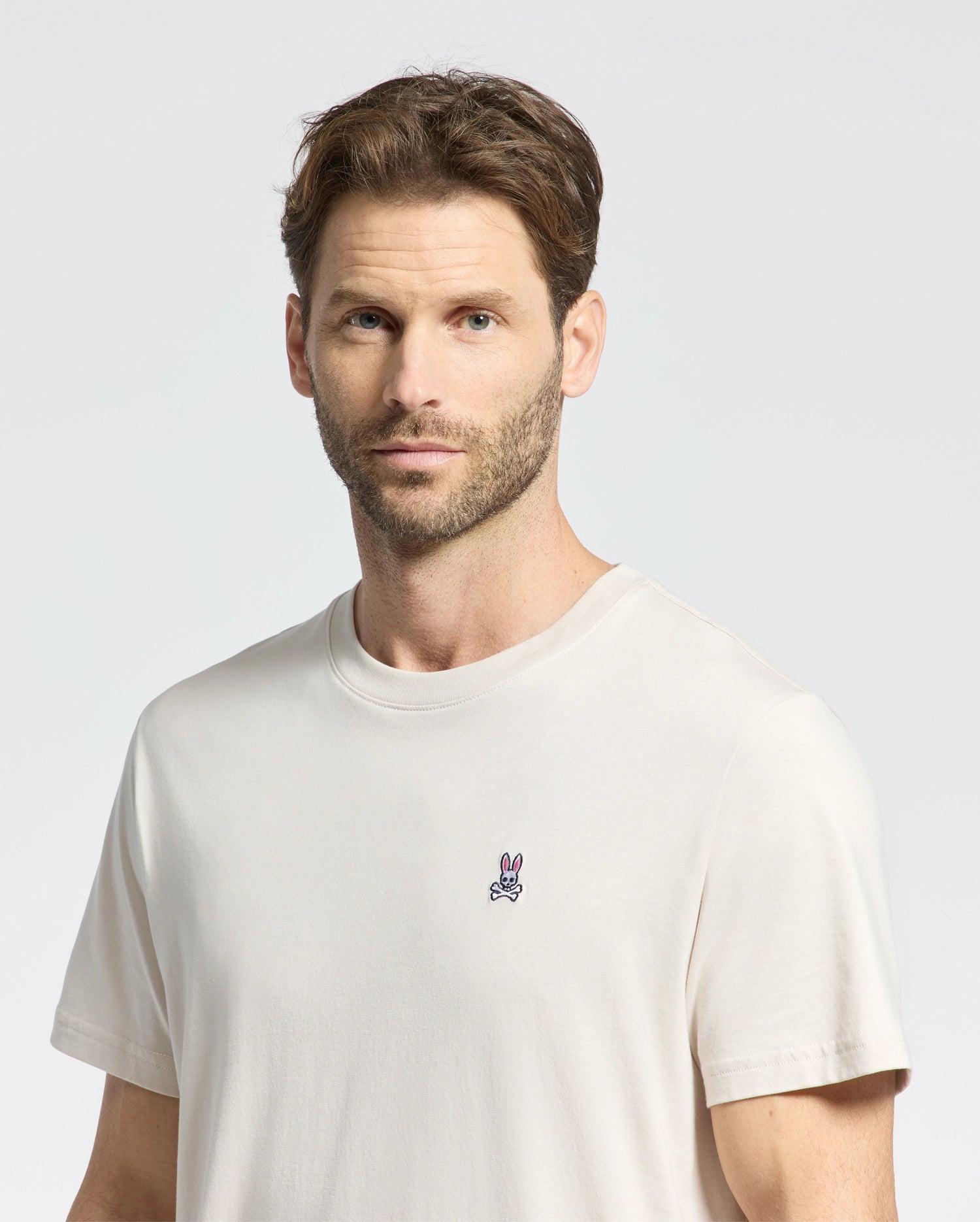 A brown-haired person with a beard wears a Psycho Bunny Men's Classic Crew Neck Tee, light beige, made from premium jersey material with an embroidered bunny logo. A plain light background emphasizes the casual Pima cotton attire.