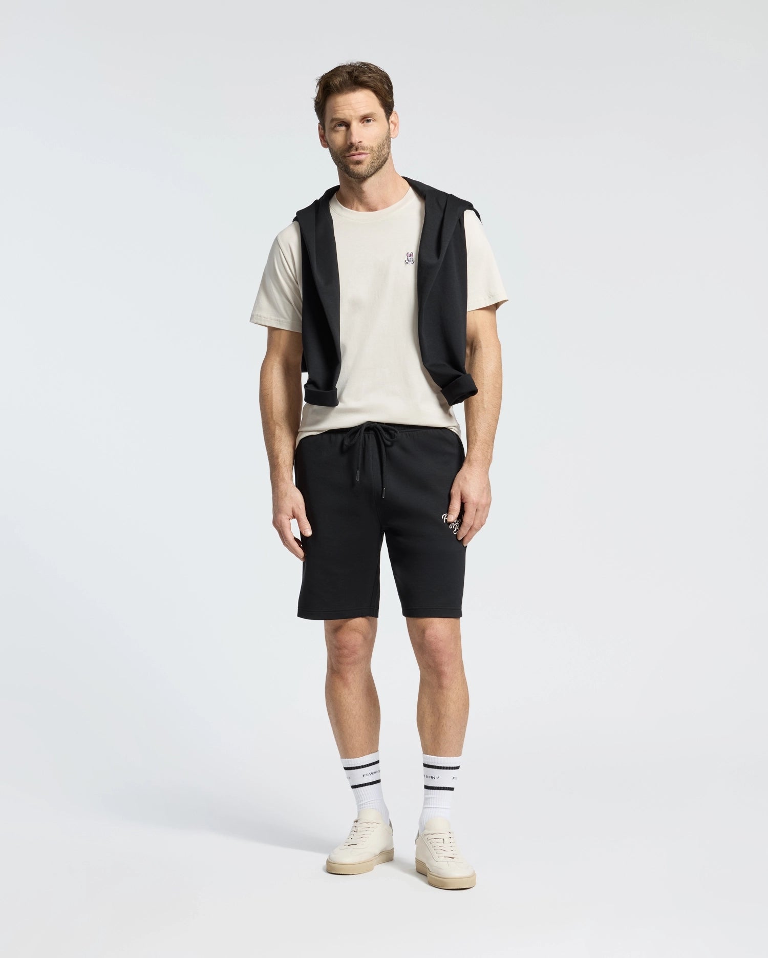 A man stands against a plain background wearing a Psycho Bunny MENS CLASSIC CREW NECK TEE (B6U014B200) in white, paired with black shorts and a black sweatshirt draped over his shoulders. His look is completed with striped socks and white sneakers.