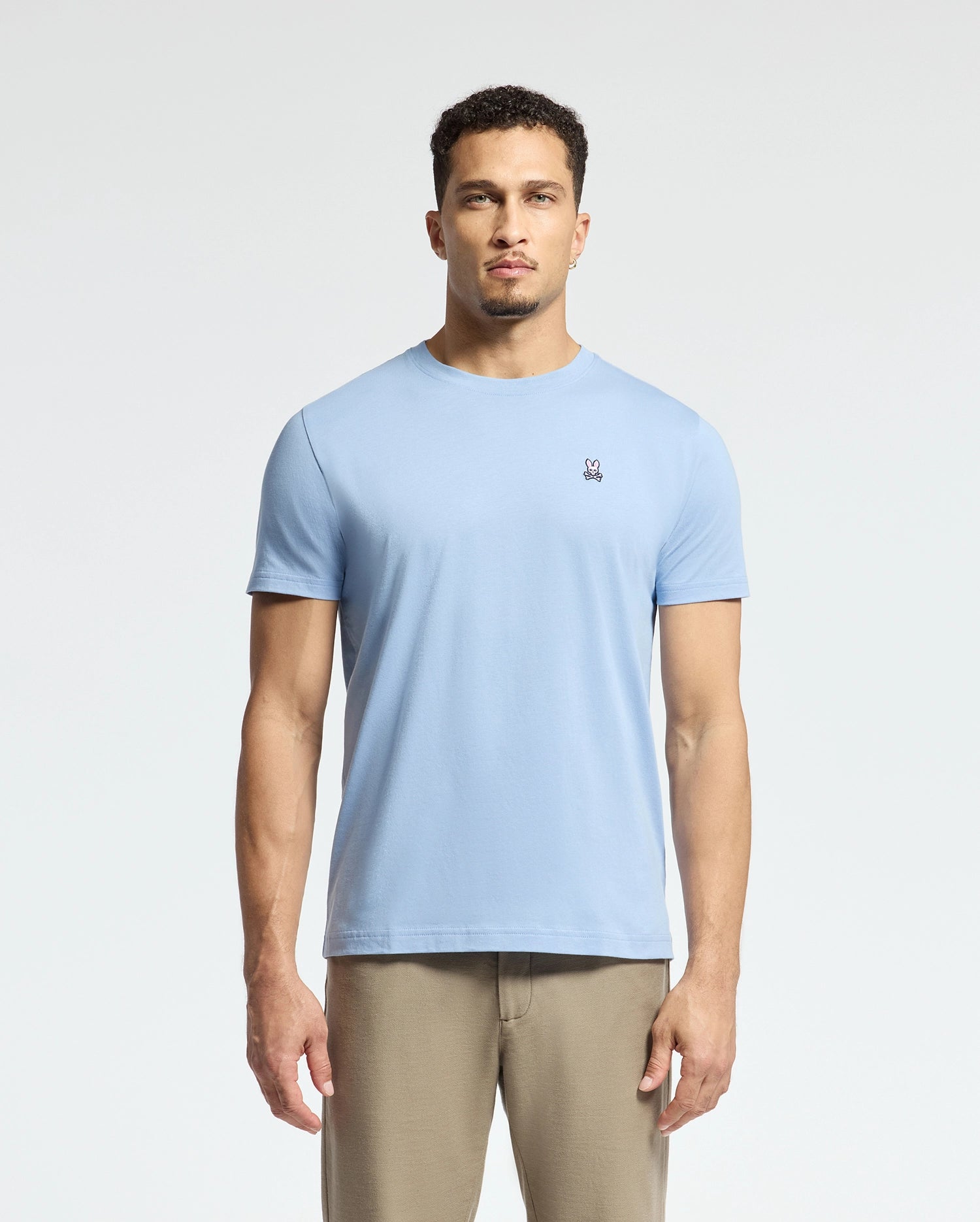 A person wearing a Psycho Bunny MENS CLASSIC CREW NECK TEE, style B6U014B200, in light blue Pima cotton with an embroidered chest logo and beige pants stands against a plain white background.