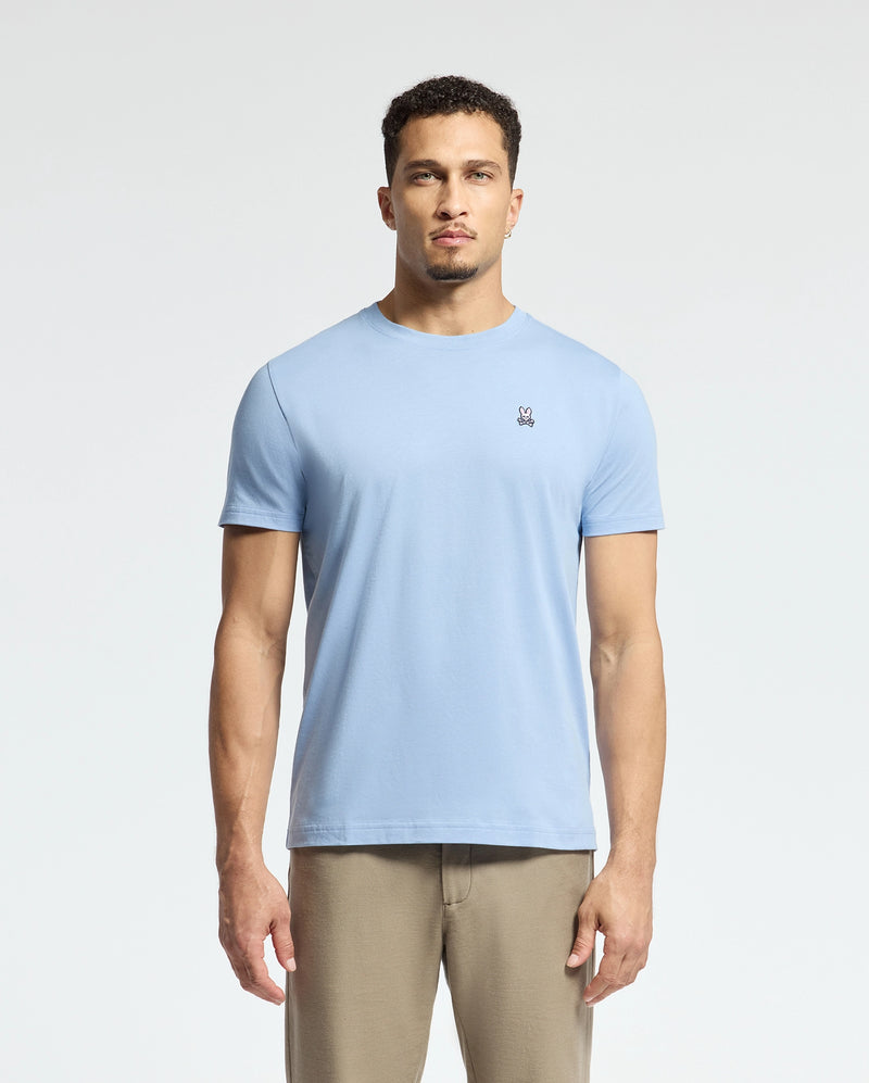 A person wearing a Psycho Bunny MENS CLASSIC CREW NECK TEE, style B6U014B200, in light blue Pima cotton with an embroidered chest logo and beige pants stands against a plain white background.
