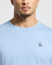 A man wearing the Psycho Bunny Men's Classic Crew Neck Tee in light blue, crafted from soft Pima cotton, featuring a small pink bunny and crossbones embroidered on the chest. His face is partially visible.