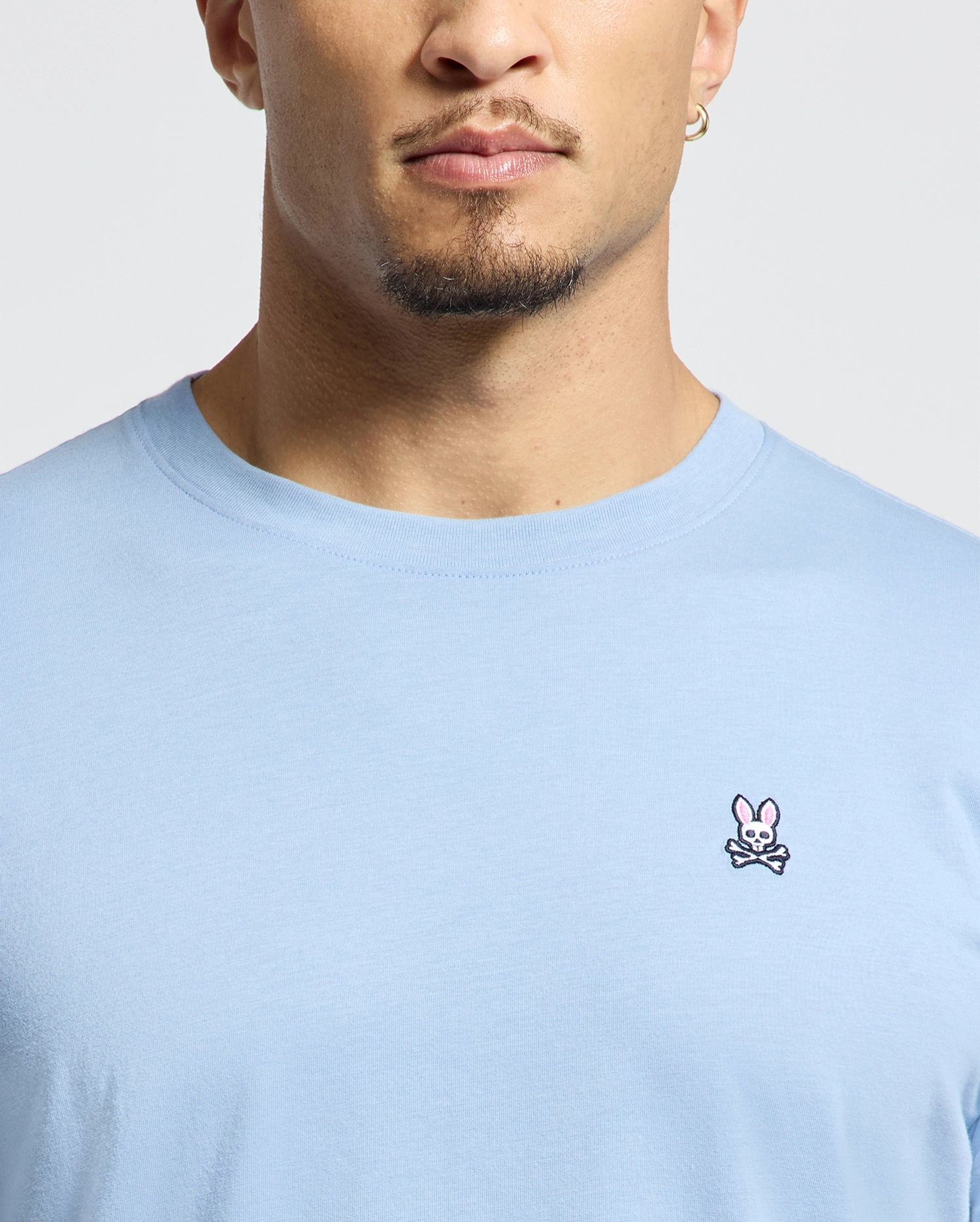 A man wearing the Psycho Bunny Men's Classic Crew Neck Tee in light blue, crafted from soft Pima cotton, featuring a small pink bunny and crossbones embroidered on the chest. His face is partially visible.