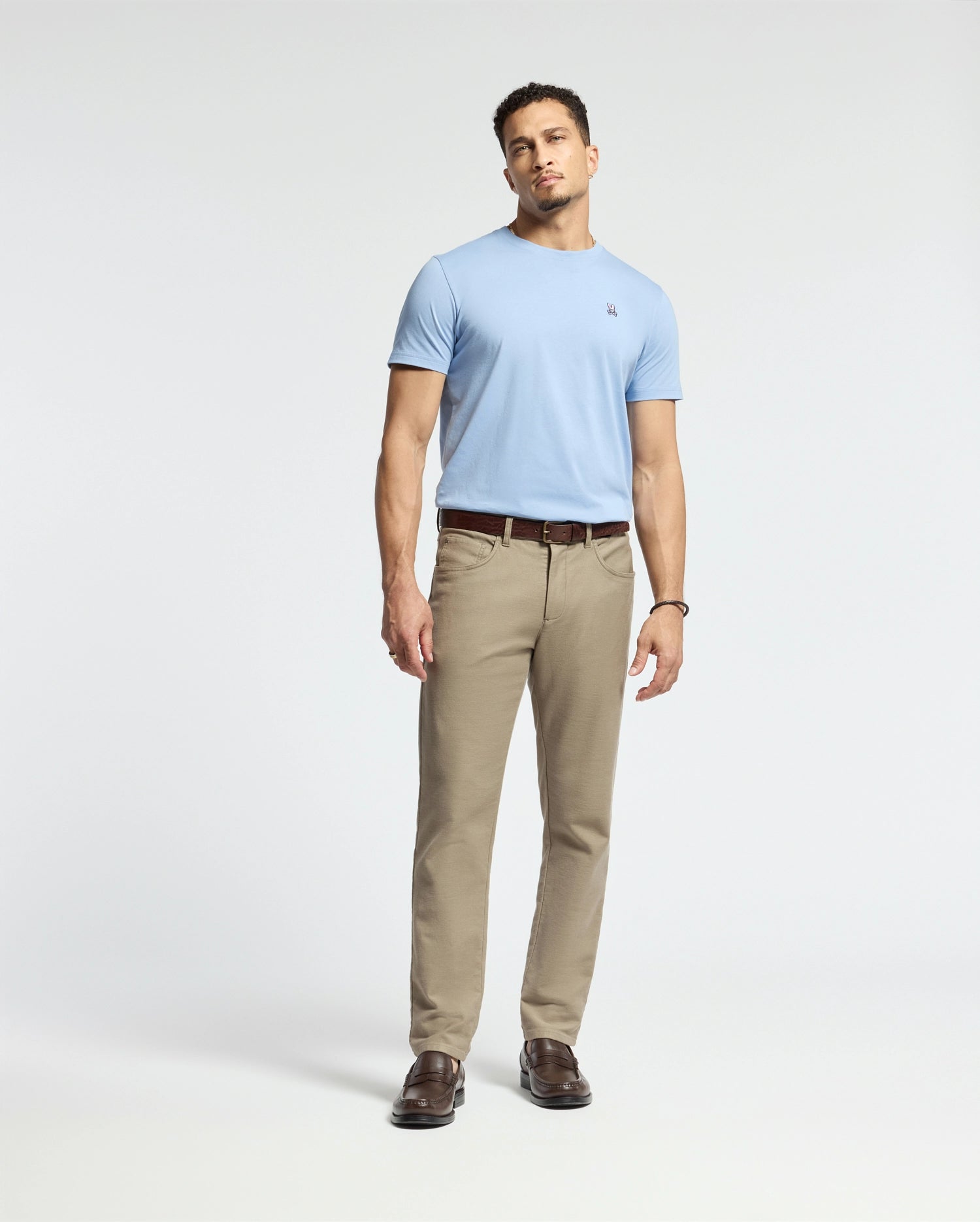 A man poses against a plain backdrop in a Psycho Bunny MENS CLASSIC CREW NECK TEE - B6U014B200, light blue, made from Pima cotton, paired with khaki pants and brown shoes. His neutral expression and relaxed stance encapsulate men's classic style.