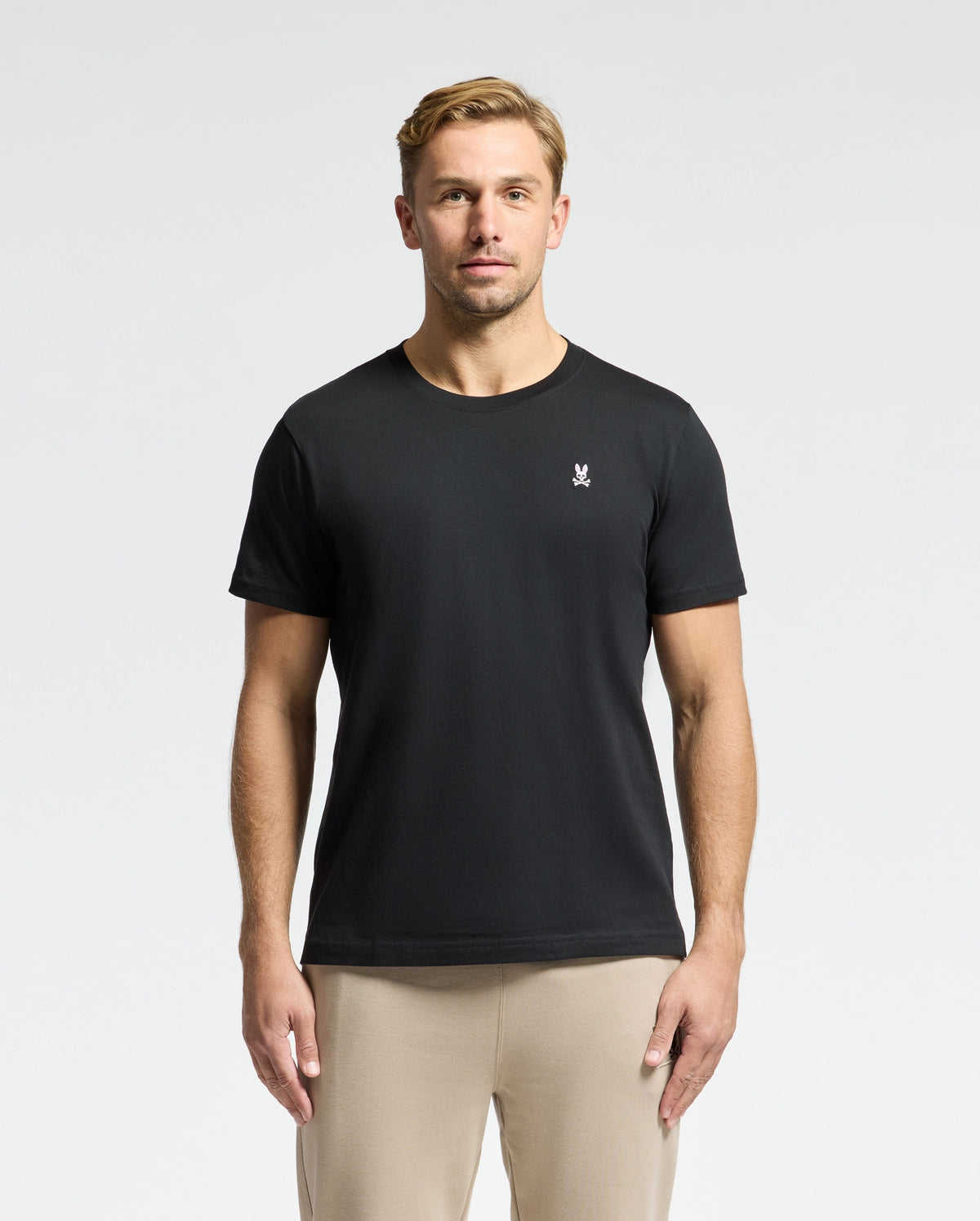 A man with short hair is wearing the Psycho Bunny MENS CLASSIC CREW NECK TEE - B6U014CRPC, crafted from 100% Pima cotton, featuring a small white chest logo. The black tee, matched with beige pants, pops against the plain white background.