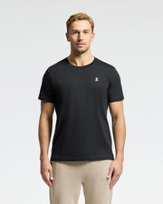 A man with short hair is wearing the Psycho Bunny MENS CLASSIC CREW NECK TEE - B6U014CRPC, crafted from 100% Pima cotton, featuring a small white chest logo. The black tee, matched with beige pants, pops against the plain white background.