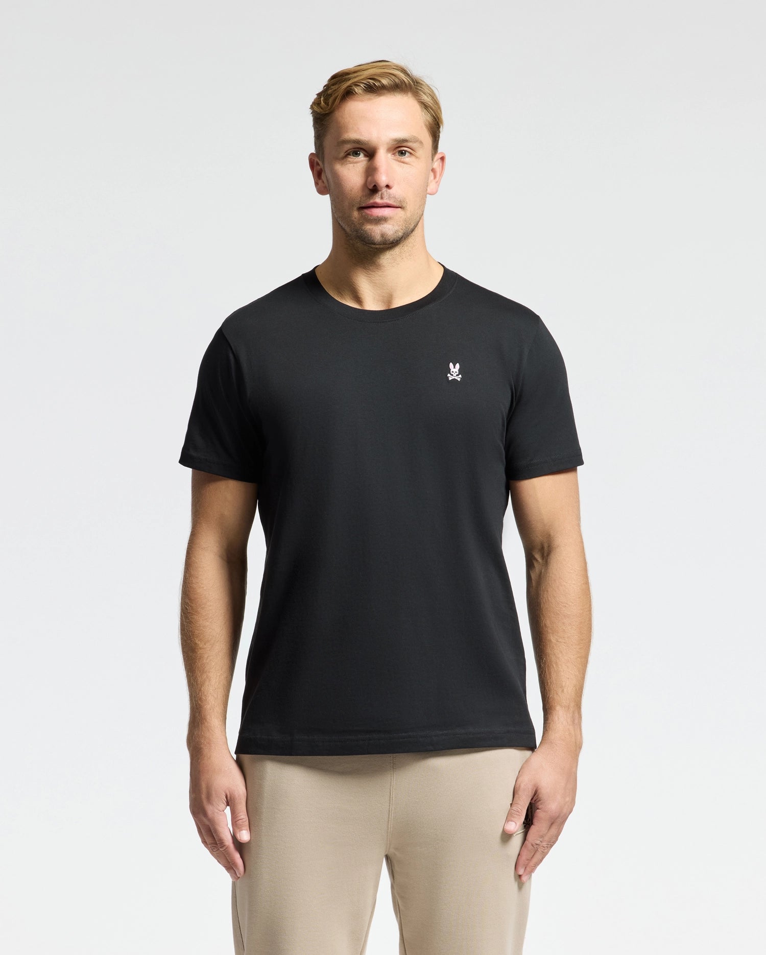 A man with short hair is wearing the Psycho Bunny MENS CLASSIC CREW NECK TEE - B6U014CRPC, crafted from 100% Pima cotton, featuring a small white chest logo. The black tee, matched with beige pants, pops against the plain white background.