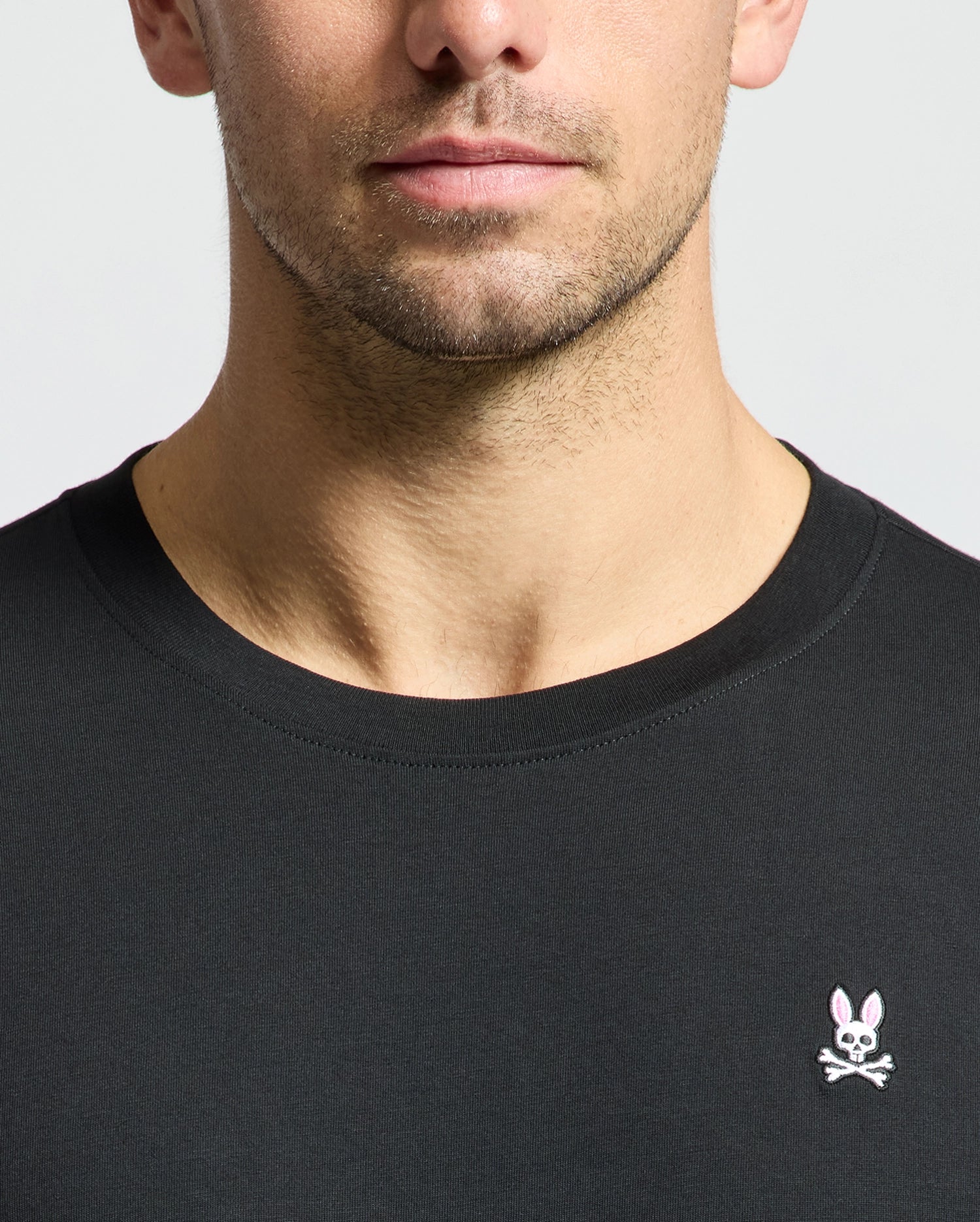 Someone is wearing a Psycho Bunny MENS CLASSIC CREW NECK TEE - B6U014CRPC in black, made from 100% Pima cotton. It has a small embroidered rabbit logo with crossed bones on the chest. The plain, light-colored background accentuates the tee's design and taped seams.