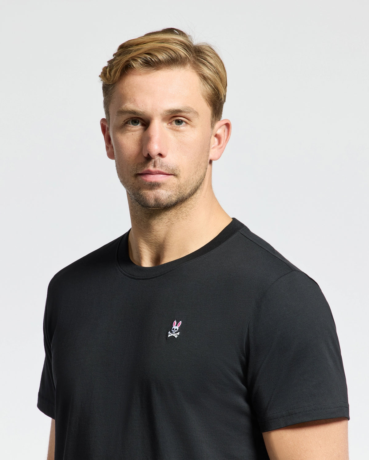 A person with short blonde hair wears a Psycho Bunny MENS CLASSIC CREW NECK TEE - B6U014CRPC, made of 100% Pima cotton, with taped seams and a small chest logo. They're set against a plain white background.