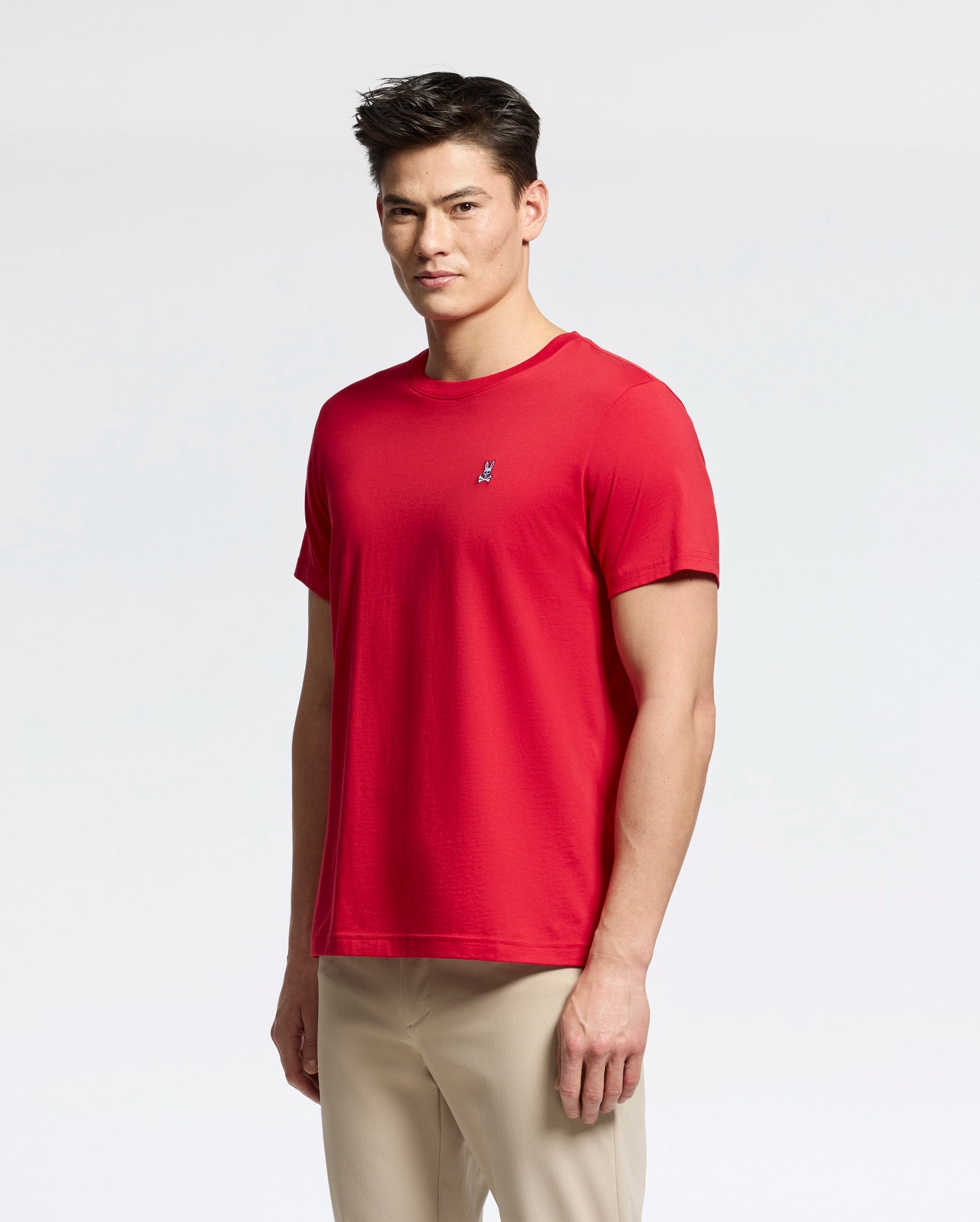 A man is wearing a Psycho Bunny MENS CLASSIC CREW NECK TEE (B6U014CRPC) in red, featuring a small embroidered logo on the chest, paired with beige pants. The classic tee is crafted from soft Pima cotton, and he stands against a plain white background.