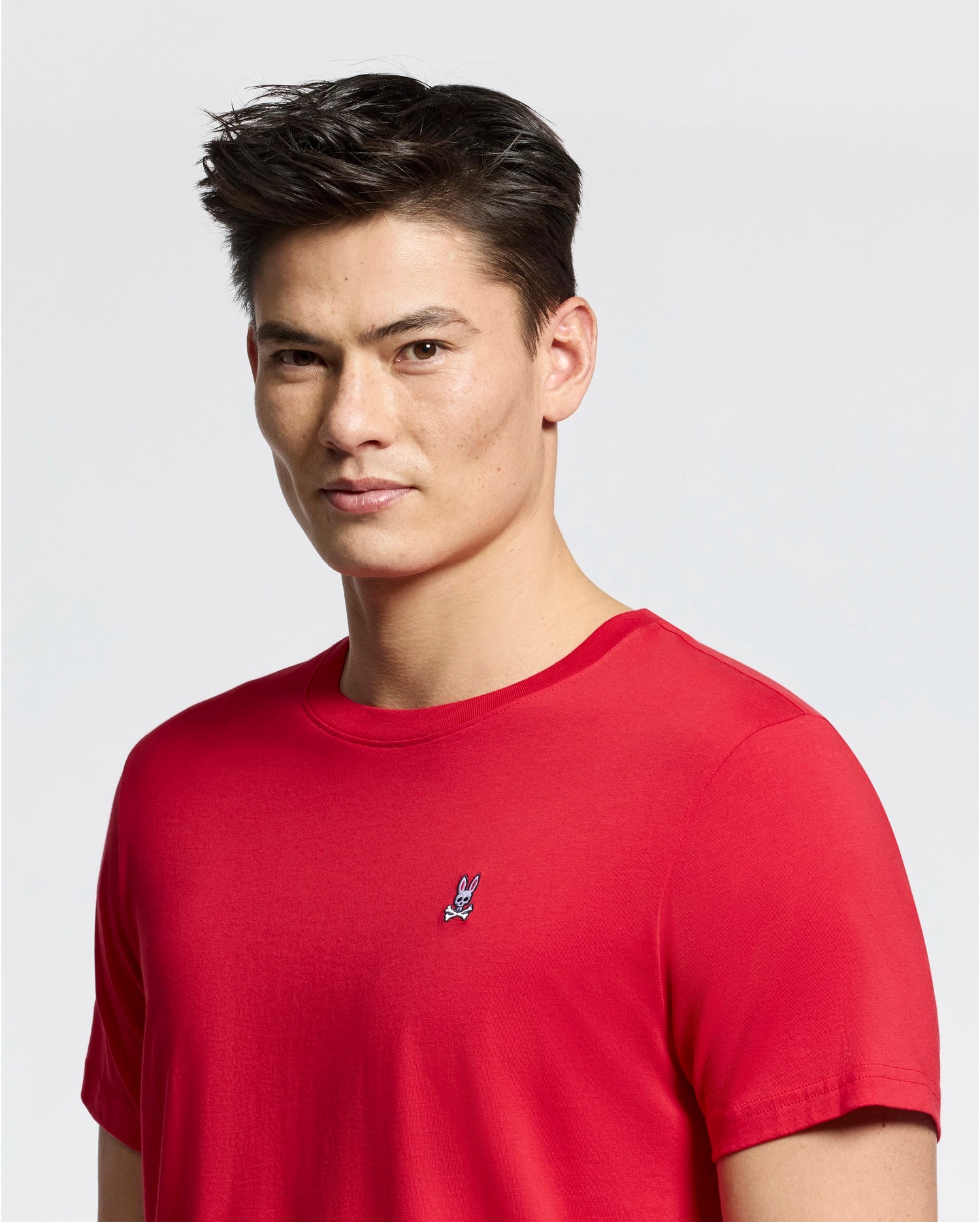 A person with short dark hair wears the Psycho Bunny MENS CLASSIC CREW NECK TEE - B6U014CRPC in red, adorned with a small embroidered rabbit logo on the chest against a plain white background.