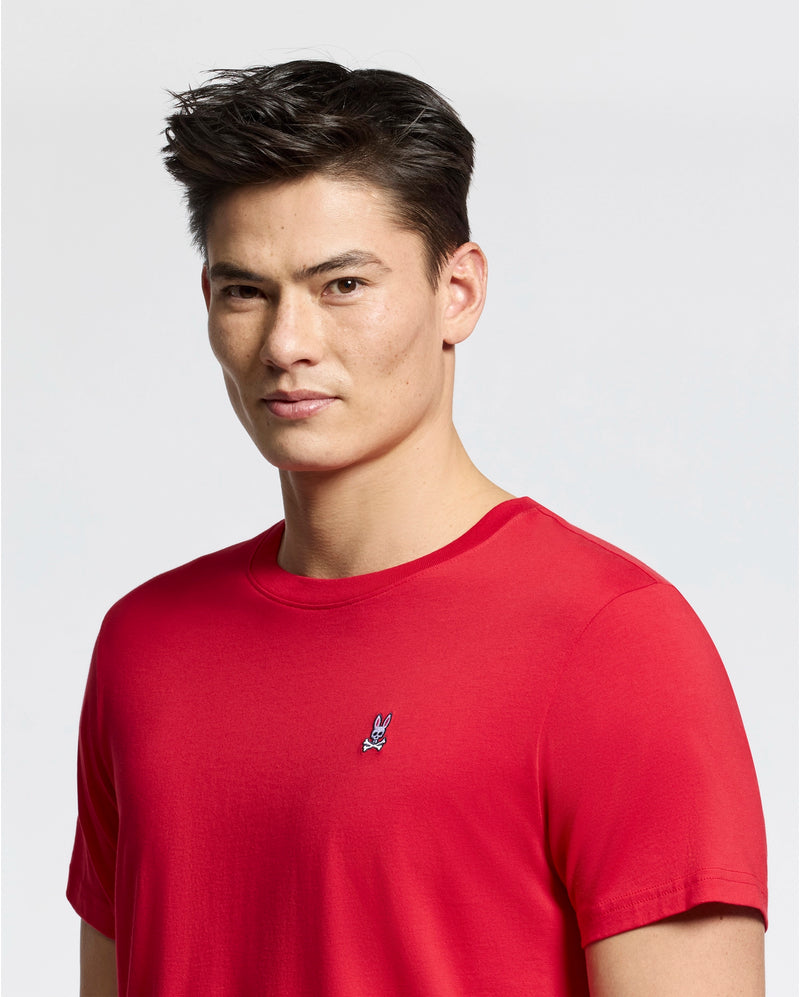 A person with short dark hair wears the Psycho Bunny MENS CLASSIC CREW NECK TEE - B6U014CRPC in red, adorned with a small embroidered rabbit logo on the chest against a plain white background.
